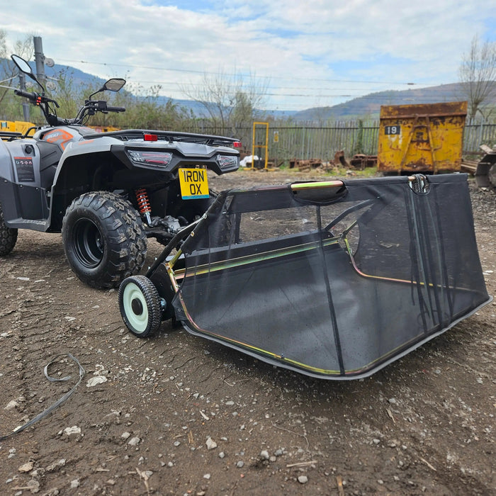 A quad bike with the license plate "IRON OX" is parked on dirt, attached to a Tow-Behind Lawn Sweeper 48" – Heavy-Duty Leaf, Grass & Debris Collector. Trees, hills, and industrial equipment appear in the background.