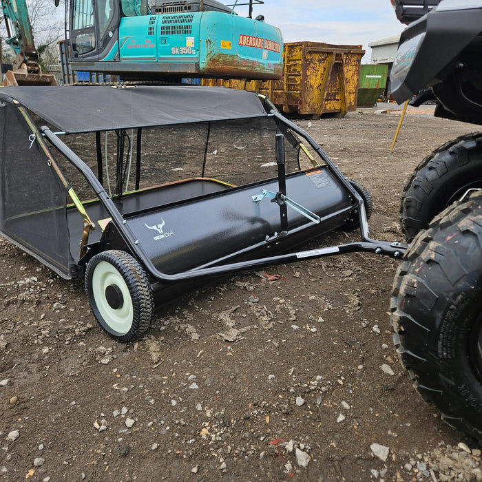 A Tow-Behind Lawn Sweeper 48" – Heavy-Duty Leaf, Grass & Debris Collector for Riding Mowers, Tractors & ATVs is attached to an ATV and parked on a gravel surface near construction equipment and dumpsters.