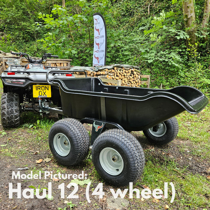 An Iron Ox® Haul 12 - Tipping Trailer 1200lb 4-Wheel with a black, corrosion-resistant bed is attached to an ATV, parked on grass with stacked firewood and trees in the background.