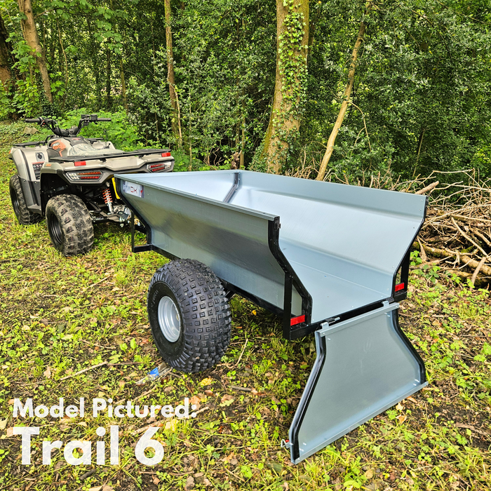 A gray All-Terrain ATV & Farm Tipping Trailer - Trail 6, with a drop-down tailgate, is attached to an ATV and parked on grassy ground in a wooded area.