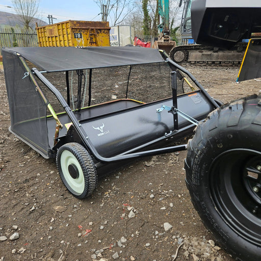 The Tow-Behind Lawn Sweeper 48" is a heavy-duty leaf, grass, and debris collector for riding mowers, tractors, and ATVs—shown here attached to a utility vehicle on gravel at an outdoor work site for efficient cleanup.