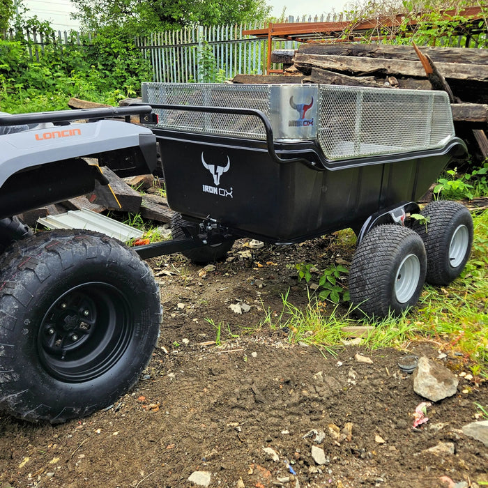 A Heavy-Duty 4 Wheel Off-Road ATV Tipping Trailer for Quad Bikes is parked on uneven ground near stacked wood and a metal fence, attached to a vehicle.
