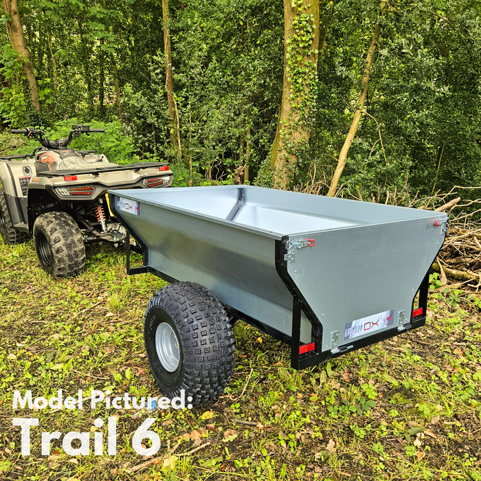 An All-Terrain ATV & Farm Tipping Trailer—Heavy-Duty Utility Trailer "Trail 6"—is attached to an ATV in a wooded area with green trees and undergrowth.