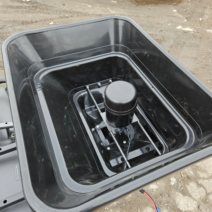 A black plastic ATV Quad Bike Seed Feed Fertiliser Spreader 12V with an electronic device installed at its base, positioned on a sandy surface with cables nearby.