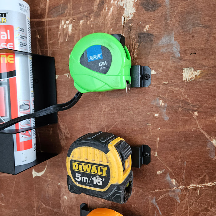 Two Tape Measure Holder - 5 Pack attached to a wooden board, one above the other; the upper one is green and labeled "5m 19mm," the lower one is yellow and black with the brand "d