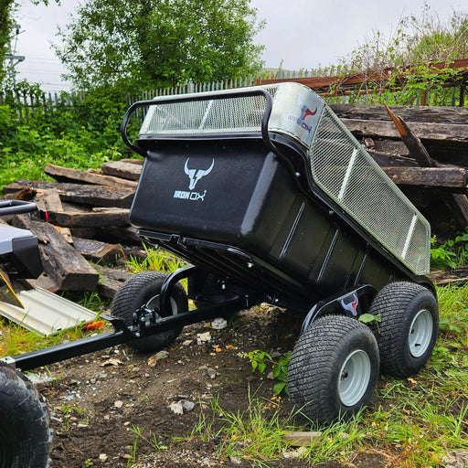 A black Heavy-Duty 4 Wheel Off-Road ATV Tipping Trailer for farming, logging, and equestrian use is parked on uneven ground near stacked wooden planks and greenery.