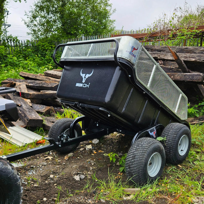 A black Heavy-Duty 4 Wheel Off-Road ATV Tipping Trailer for farming, logging, and equestrian use is parked on uneven ground near stacked wooden planks and greenery.