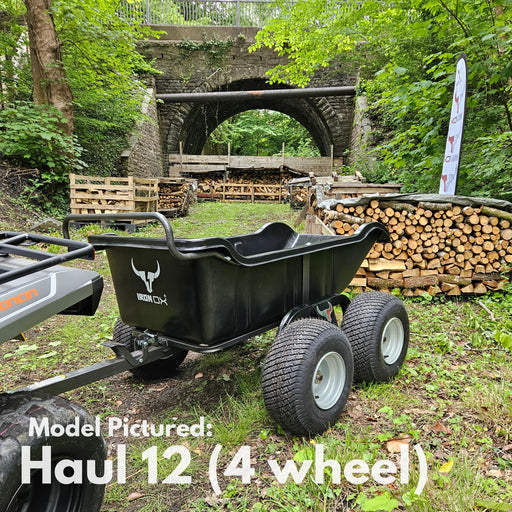 An Iron Ox® Haul 12 - Tipping Trailer 1200lb 4-Wheel in black, with a corrosion-resistant bed, is attached to a vehicle in a wooded area, stacked with firewood and set against a stone archway backdrop.